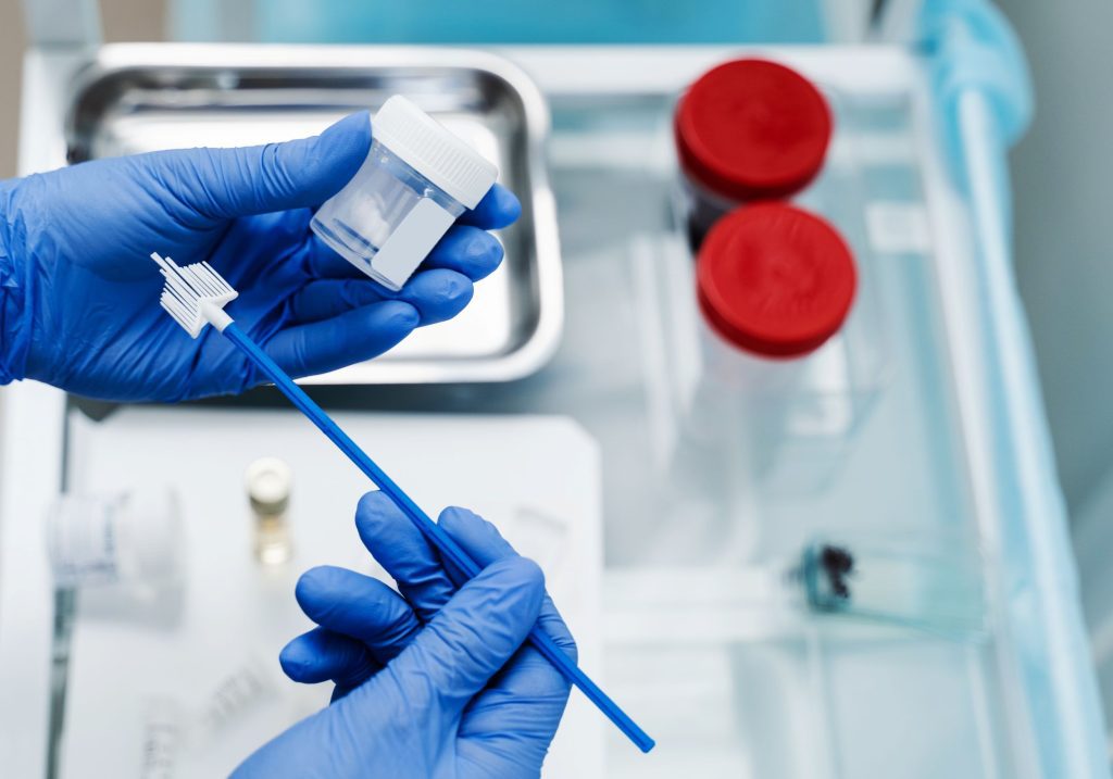 Położna Centrum Medycznego neoMedica w Poznaniu trzyma w rękawiczkach lekarskich szczoteczkę do cytologii płynnej, a w tle widoczne są zamazane pojemniki.
