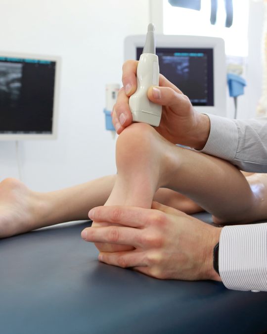 Głowica USG przykładana do kostki małego Pacjenta, a w tle zamazany sprzęt USG - ilustracja dotycząca USG narządów ruchu u dzieci w Centrum Medycznym neoMedica w Poznaniu.